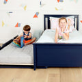 Two children in a bright kids' bedroom with a navy blue twin bed and pull-out trundle mattress; one child reading a picture book while the other smiles on pastel bedding against geometric wallpaper.