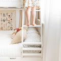 Child standing barefoot on a white bunk bed ladder in a bright nursery with floral wall art and cozy pillows