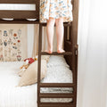 Child standing barefoot on wooden bunk bed ladder in cozy kids bedroom with patterned dress, gingham pillows and botanical wall art