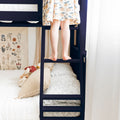 Child standing barefoot on ladder of navy bunk bed in a cozy kids bedroom with patterned dress, floral wall art and decorative pillows