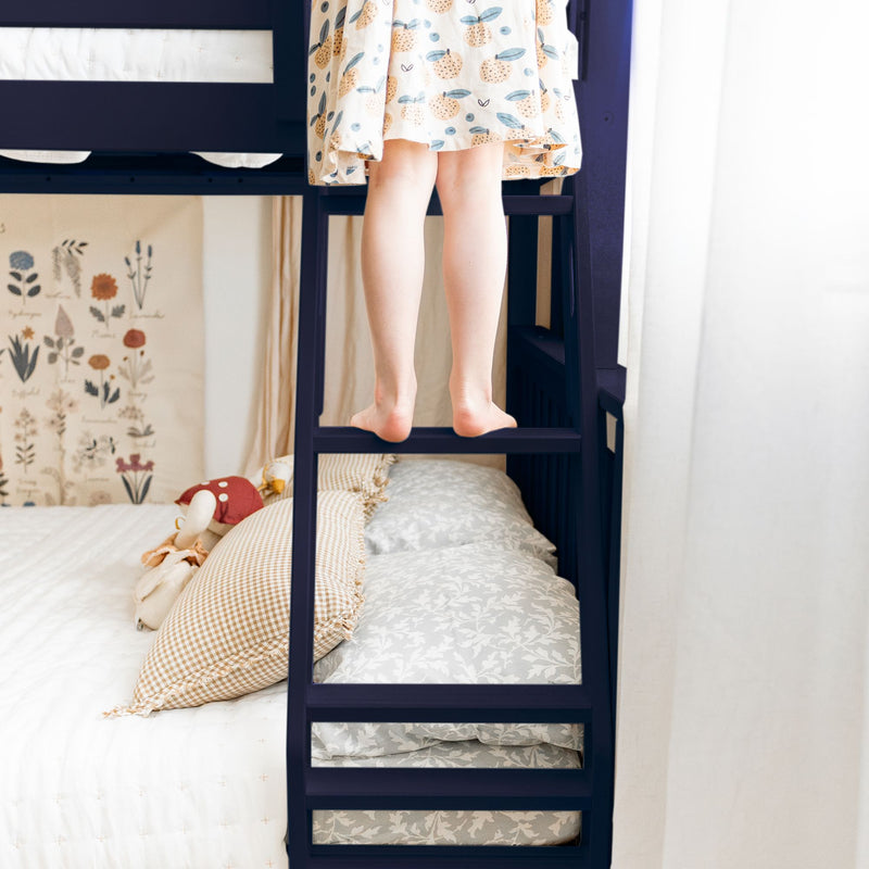 Child standing barefoot on ladder of navy bunk bed in a cozy kids bedroom with patterned dress, floral wall art and decorative pillows