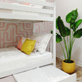 White wooden bunk bed with pull-out trundle, crisp white bedding and yellow pom-pom pillow against pink geometric wallpaper and potted banana-leaf plant — modern kids' bedroom decor