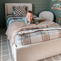 Smiling young boy lying on a cozy bed in a modern children's bedroom with plaid bedding, soft throw blanket, and neutral decor
