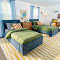 Shared kids' bedroom with two blue upholstered twin beds, green comforters, orange checkered pillows, wooden nightstand, yellow-striped rug and playful wall decor