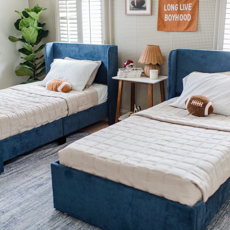 Boys' bedroom with twin navy-blue upholstered beds, beige quilts, football plush, wicker bedside lamp, fiddle-leaf fig and shuttered windows