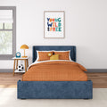 Children's bedroom with blue upholstered bed, orange quilted bedding, framed "Young Wild and Free" wall art, white nightstand with orange lamp and blue alarm clock, and soccer ball on wooden floor