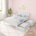 Cozy girls bedroom with white wooden twin bed, pastel pillows and throw, pink scallop wallpaper, framed rainbow and sun prints, blue bedside lamp, potted plant and under-bed storage drawers