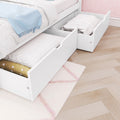 White bed with underbed storage drawers filled with quilts and pillows, blue throw blanket, pink patterned wallpaper and light wood herringbone floor