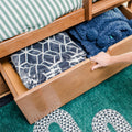 Under-bed storage drawer in wooden bed frame opened by hand revealing folded fleece and patterned blankets with a green-striped mattress sheet for organized bedding storage
