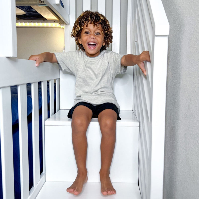 Smiling child with curly hair sitting on a white bunk bed ladder in a bright kids' bedroom, wearing a gray t-shirt and black shorts