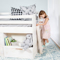 White wooden loft bunk bed with patterned pillows, stuffed bunny, children's books and a young girl climbing the ladder on a blue rug