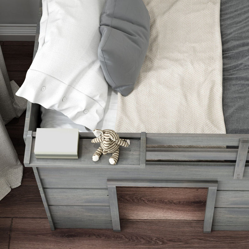 Top view of gray wooden toddler bed with built-in shelf holding a striped plush elephant toy and book, white and gray pillows and beige blanket on hardwood floor