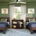 Cozy kids bedroom with two twin wooden beds, green paneled walls, cowboy-themed framed art, ceiling fan, sheer curtains, rattan bedside lamp, alarm clock and stuffed cow toy