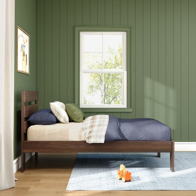Sunlit kids' bedroom with green vertical paneled walls, white-trimmed window, wooden single bed with navy and plaid bedding and green pillows, light blue rug and wooden toy on the floor
