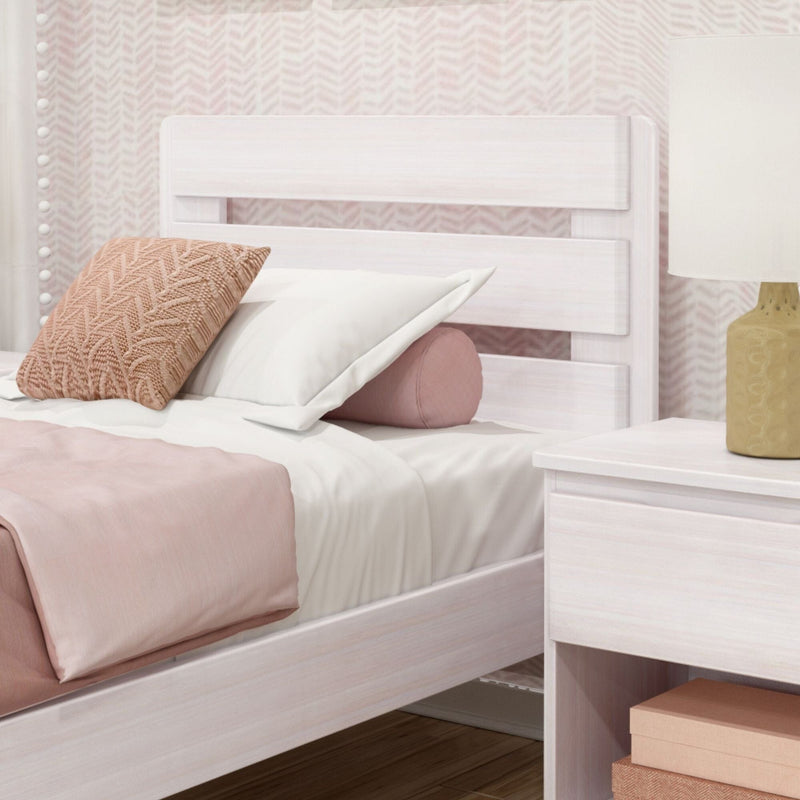 Whitewashed wooden bed frame with textured pink throw pillow, round pink bolster and white linens beside a nightstand with gold lamp