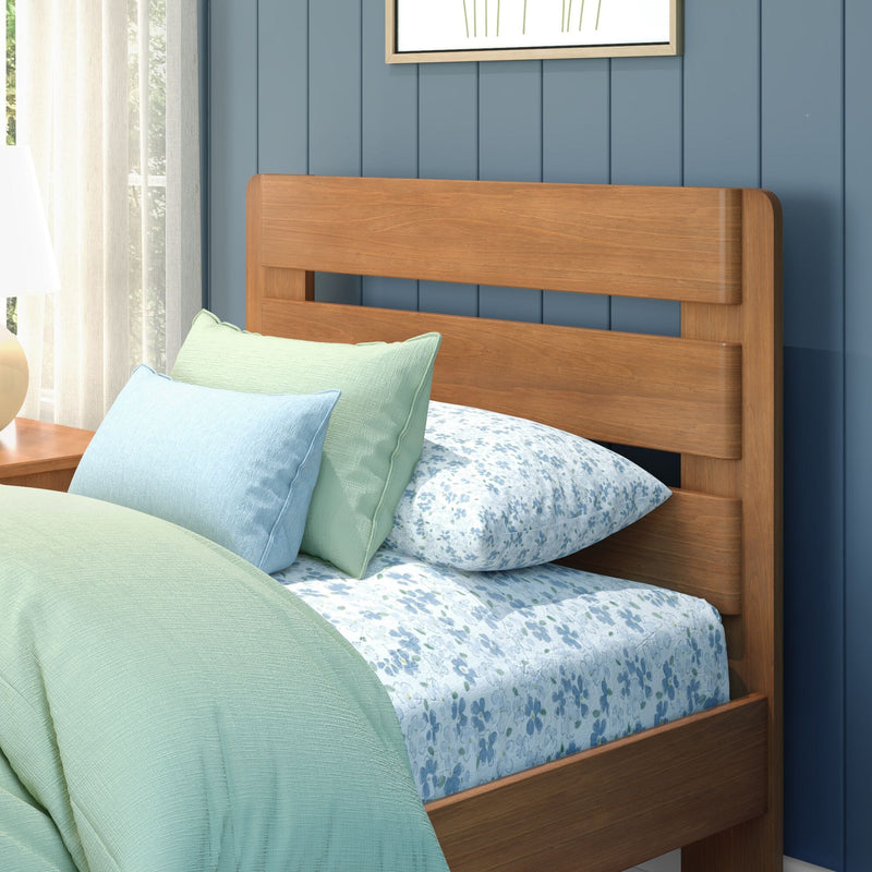Modern wooden bed headboard against blue paneled wall with mint green and light blue pillows, floral bedding, bedside lamp and sheer curtains