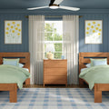 Cozy twin bedroom with wooden twin beds and green bedding, central dresser and lamp, sheer curtains over a sunlit window, navy blue paneled accent wall with yellow floral art and ceiling fan