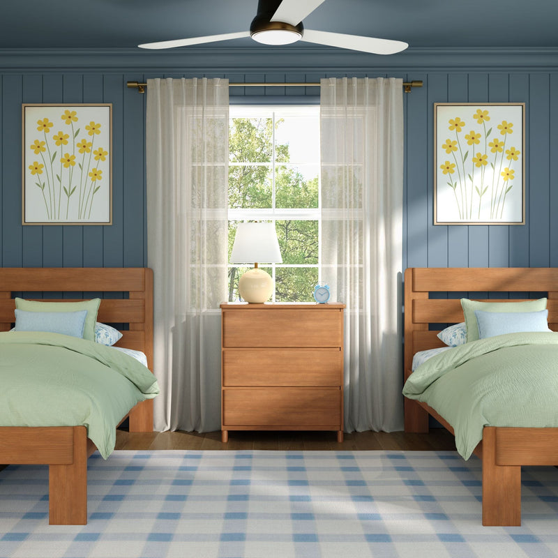 Cozy twin bedroom with wooden twin beds and green bedding, central dresser and lamp, sheer curtains over a sunlit window, navy blue paneled accent wall with yellow floral art and ceiling fan