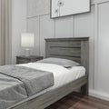 Modern farmhouse bedroom with rustic gray wood headboard and matching nightstand, white bedding with gray throw blanket, ceramic table lamp, neutral curtains and board-and-batten wall with framed botanical art