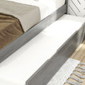 Modern grey wood platform bed with under-bed pull-out trundle drawer, white mattress and beige linen bedding on hardwood floor with white tasseled rug — space-saving bedroom storage solution