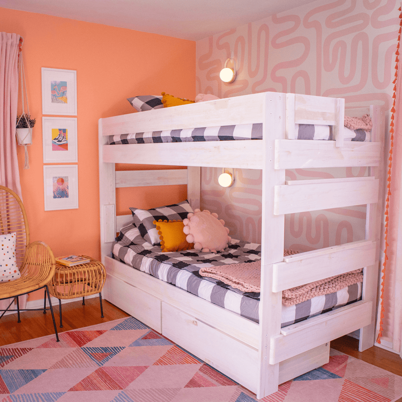 Pastel peach and pink kids' bedroom interior with white wooden bunk bed, black-and-white checkered bedding, pom-pom pillows, rattan chairs and geometric rug