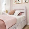 Pastel pink bedroom with white wooden twin bed and trundle, blush knit throw pillow, white bedside lamp, floral wall art and natural window light