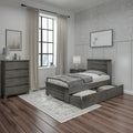 Modern farmhouse bedroom featuring a grey wood twin bed with under-bed storage drawers, matching dresser and nightstand, neutral bedding, linen curtains, black-framed window, floral wallpaper accent, and dark hardwood floors.