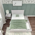 Overhead view of a cozy modern farmhouse bedroom featuring a whitewashed wooden twin bed with green duvet and pillows, sage green board-and-batten accent wall topped with botanical wallpaper, bedside table with lamp, trundle drawer and dark hardwood flooring.