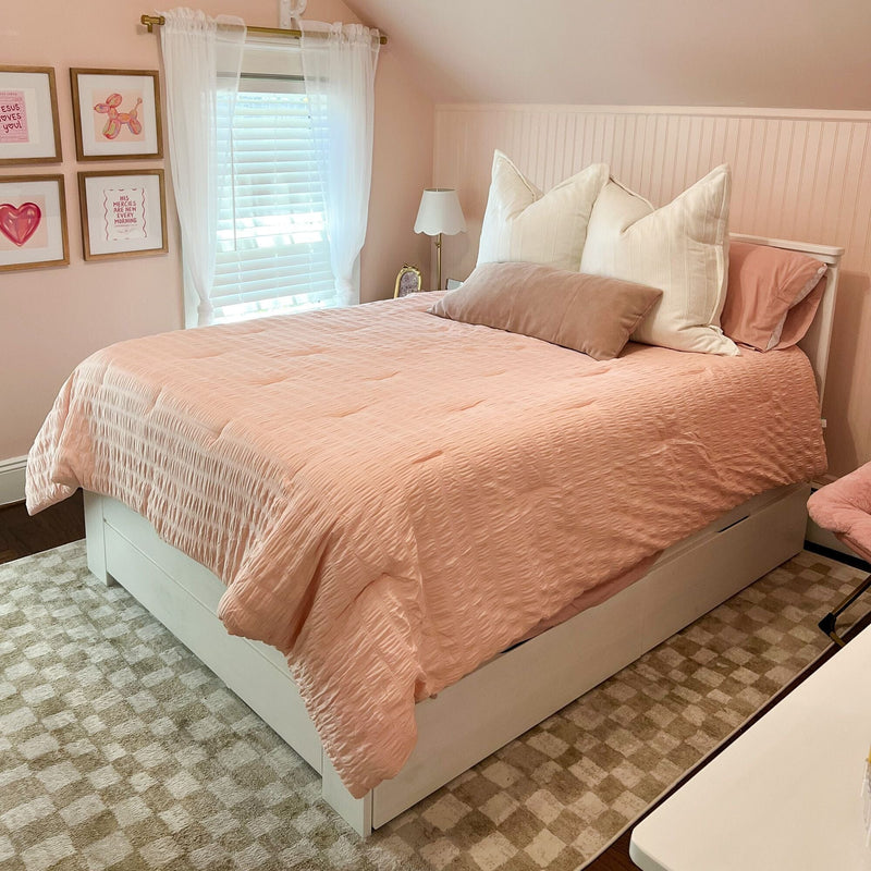 Cozy blush-pink bedroom with quilted pink comforter, white oversized pillows on a white storage bed frame, sheer curtains over white plantation shutters, pink beadboard accent wall, framed gallery art and brass-accent bedside lamp