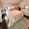 Cozy pastel pink bedroom with blush quilted bedding and pillows, wooden nightstand, white dresser with round mirror, pink accent chair, potted plant and checkered rug.