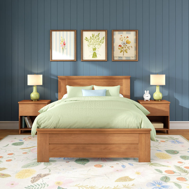 Modern farmhouse bedroom with wooden bed frame and green bedding, blue paneled wall with three framed floral prints, matching green bedside lamps, wooden nightstands and floral rug