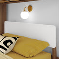 Modern brass globe wall sconce above bed with yellow bedding and black-and-white accent pillow on wooden bed frame, bedroom wall lighting