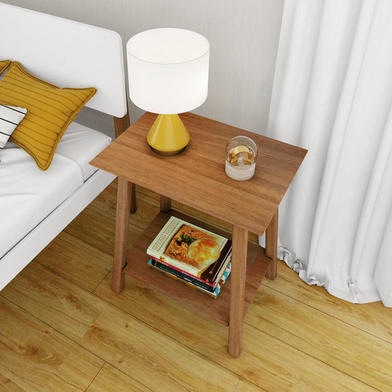 Wooden bedside table nightstand with yellow lamp, glass ornament and stacked books next to white bed with mustard pillow and white curtains on hardwood floor.