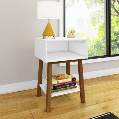 Modern white nightstand with wooden legs and open cubby, lower shelf stacked with books, yellow table lamp and glass orb on top, by a large window overlooking a city park on hardwood floor