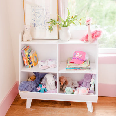 White cubby bookshelf in a nursery with pastel plush toys, children's books, a pink cap and a vase of flowers