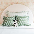 Nursery bed with round beige headboard, floral wallpaper backdrop, green sunburst-pattern pillows, striped ruffle pillow and plush toy