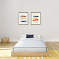 Minimalist kids' bedroom with twin bed, light blue bedding and navy pillow, neutral gray wall, two modern abstract framed prints, rattan basket and yellow area rug.