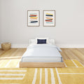Minimalist children's bedroom with light wood platform bed, blue pillow, two abstract framed prints, wicker basket and yellow geometric rug.