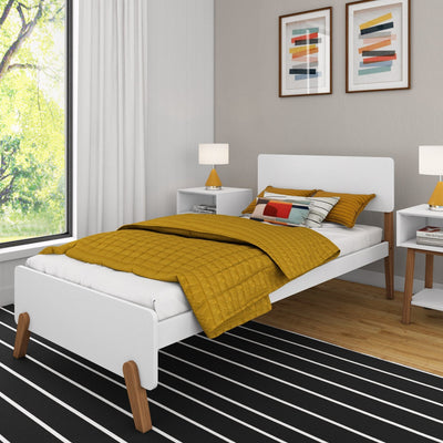 Modern minimalist bedroom with mustard yellow quilted bedding on a white platform bed, white bedside tables with yellow table lamps, abstract framed wall art, black-and-white striped rug and large window with leafy green view