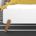 Close-up of modern white platform bed corner with mustard yellow quilt, angled wooden legs and black-and-white striped rug in minimalist bedroom interior