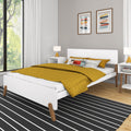 Modern minimalist bedroom with mustard yellow quilted bedding on white platform bed, bedside lamp and nightstand, abstract wall art, large window and black striped rug