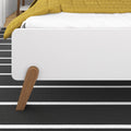 Close-up of modern white platform bed footboard with mustard yellow quilt, wooden bed leg and black-and-white striped rug in minimalist bedroom