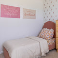 Girls bedroom with neutral beige duvet, floral pillowcase and blush pink upholstered headboard, pink and cream wall banners reading "wildflower" and "girlhood" and scattered flower wall decals — feminine bedroom decor inspiration