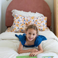 Young girl reading a picture book on a white bed with floral pillows and a pink upholstered headboard