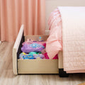 Under-bed storage drawer filled with colorful dress-up costumes beneath a tufted pink bed and soft pink quilt in a girl's bedroom with matching pink curtains