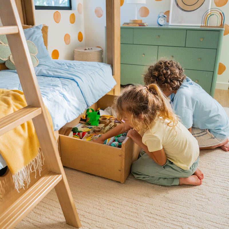 Children playing with toys at an open under-bed storage drawer in a bright kids' bedroom with bunk bed, blue bedding, green dresser and polka-dot wall