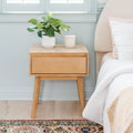 Light wood mid-century bedside table with drawer beside upholstered bed, potted green plant and candle on top, pale blue wall and patterned area rug, modern minimalist bedroom decor
