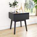Modern black nightstand bedside table in mid-century modern style with single drawer and tapered legs, styled with white vase of greenery, wooden tripod lamp and gold clock on hardwood floor by a window with forest view