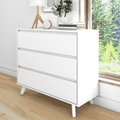 White modern three-drawer dresser with angled wooden legs in a minimalist bedroom, styled with a table lamp, clock, stacked books and plant by a sunny window