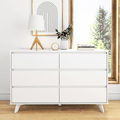Minimalist white dresser styled with a modern wooden tripod lamp, gold-accent clock, white vase of green foliage, geometric framed art and stacked books by a sunlit window — Scandinavian interior decor